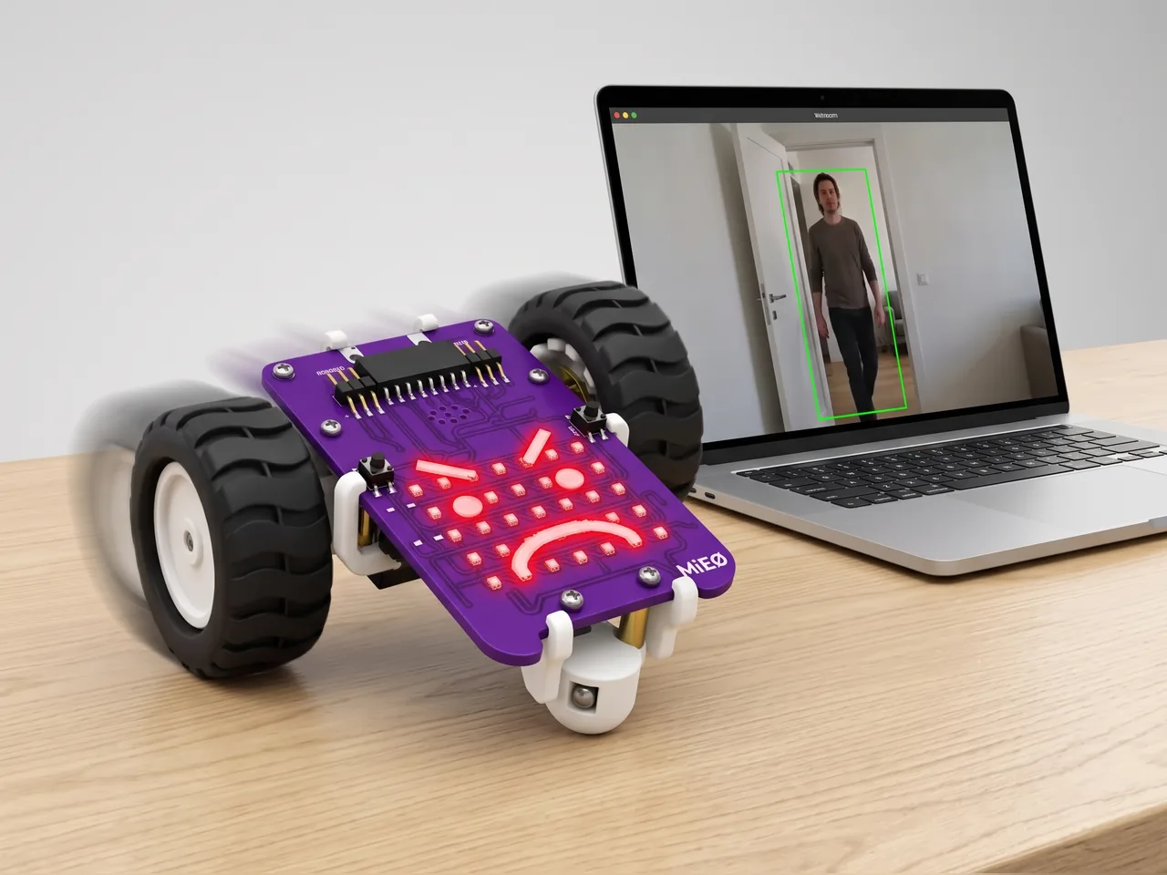 Two-wheel MIEO with an angry face on its LED grid lunging forward beside a laptop whose webcam has spotted a person